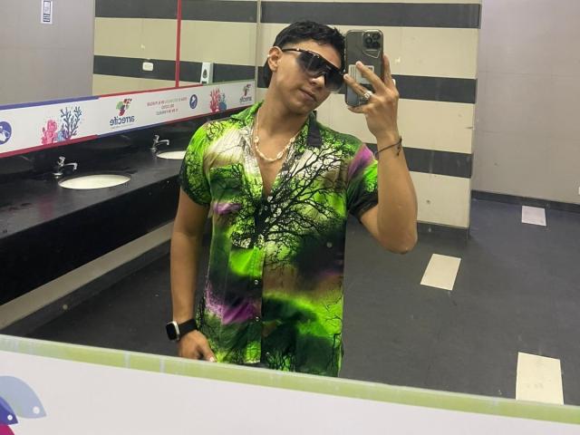 RichiMontes, com cabelo preto curto, usando óculos de sol, uma camiseta tie-dye verde e roxa, um relógio preto e um colar, tirando uma selfie no espelho em um banheiro..