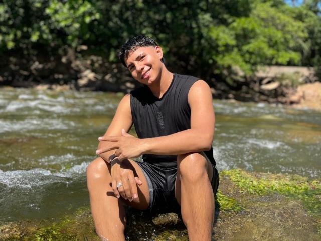 RichiMontes, vestindo uma camisa preta sem mangas e shorts pretos, sentado em uma rocha coberta de musgo em um riacho raso, sorrindo, com as mãos nos joelhos, a luz do sol destacando seu corpo suado, musculoso e bronzeado..