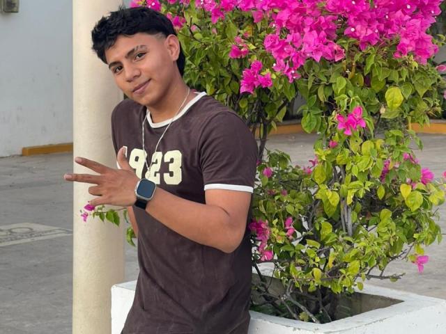 RichiMontes, un hombre de piel morena con cabello negro rizado, que lleva una camisa marrón y un reloj negro, está de pie junto a una buganvilla rosa, mostrando un gesto de paz con una sonrisa relajada..