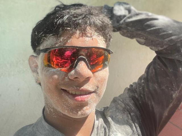 RichiMontes, com cabelo preto e suado, camisa cinza e óculos de sol com tom alaranjado, coberto de pó branco de Holi, sorrindo, com a mão direita levantada em direção à cabeça..