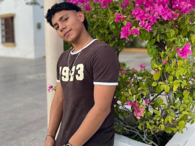 RichiMontes, un joven con cabello negro rizado, que lleva una camiseta marrón con la inscripción "1973", está de pie frente a una buganvilla rosa, mirando hacia abajo con una ligera sonrisa. Tiene las manos en los bolsillos, con una expresión relajada..