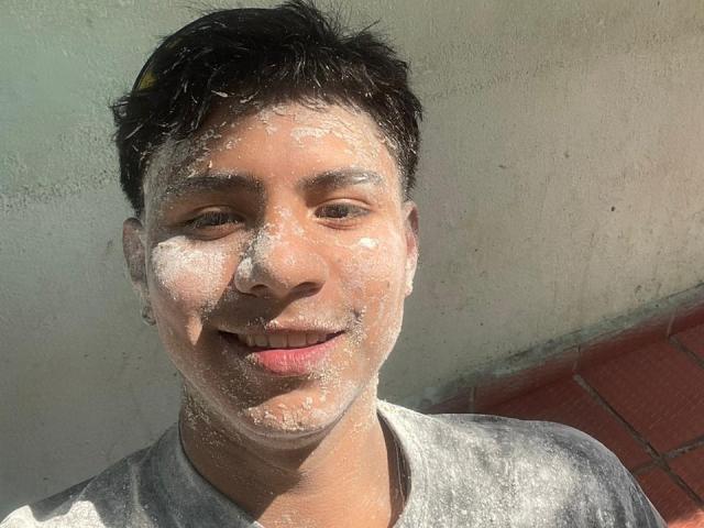 RichiMontes, um homem asiático suado com cabelo preto curto, tem pó branco por todo o rosto e na camisa cinza, sorrindo levemente para a câmera. A luz do sol realça sua aparência desgrenhada..