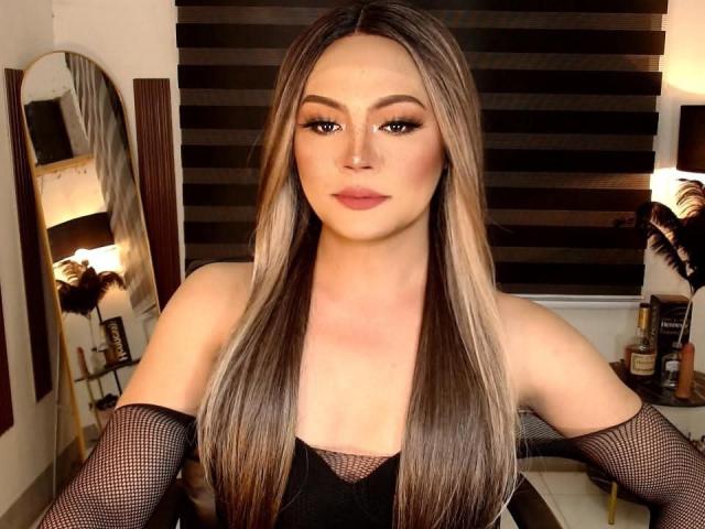 QueenAmaraTS has long, straight brown hair with blonde highlights, wearing black fishnet sleeves and a black top, looking directly at the camera with a neutral expression.