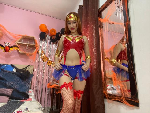 SidneyRoberts in Wonder Woman cosplay, wearing a gold headband, red vinyl bra, blue micro-skirt, fishnet stockings, red garters, and a red thong, standing in a Halloween-themed room.