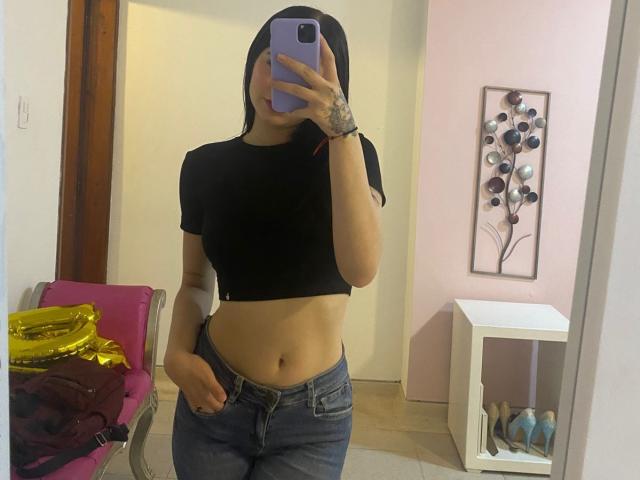 AishaBell with black crop top, showing her midriff, blue jeans, tattoo on hand, phone covering face, standing in room, mirror selfie, visible belly button, hand in pocket.