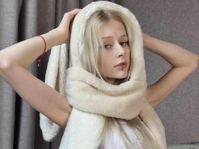 ExcitedBlizzard has blonde hair, wearing a fluffy white scarf, hands in her hair, with a red bracelet on her left wrist, looking to the side, pale skin, and freckles.