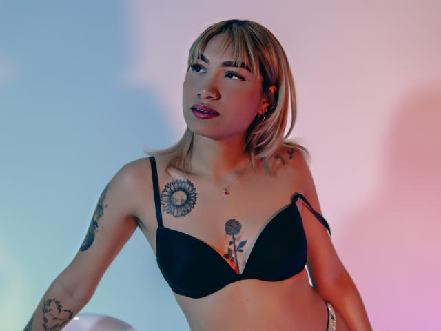 AnneGela with blonde bob, black bra, tattoos including sunflower and flower on chest, looking up, pink and blue light on her. Boobs in bra, arms raised slightly.