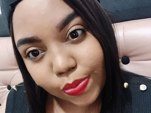 SarahSmity's face, with dark brown eyes, red lipstick, and black hair, making a pouty expression, sitting in a pink leather chair with black buttons.