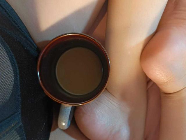A mão de KariCarter segura uma pequena xícara marrom com um líquido escuro, apoiada em seu pé descalço e na borda de uma roupa íntima de malha preta. Seus dedos estão ligeiramente curvados..
