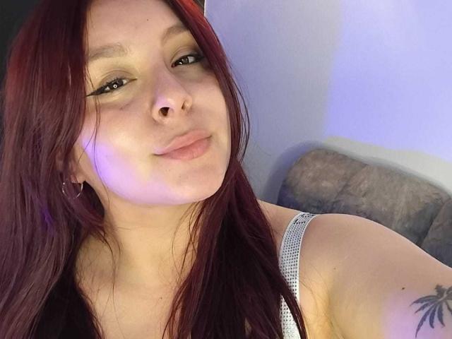 EmaJames tiene el pelo rojo, piel clara y un tatuaje en su brazo derecho. Lleva una camiseta blanca y tiene labios pequeños y finos. Está mirando a la cámara..