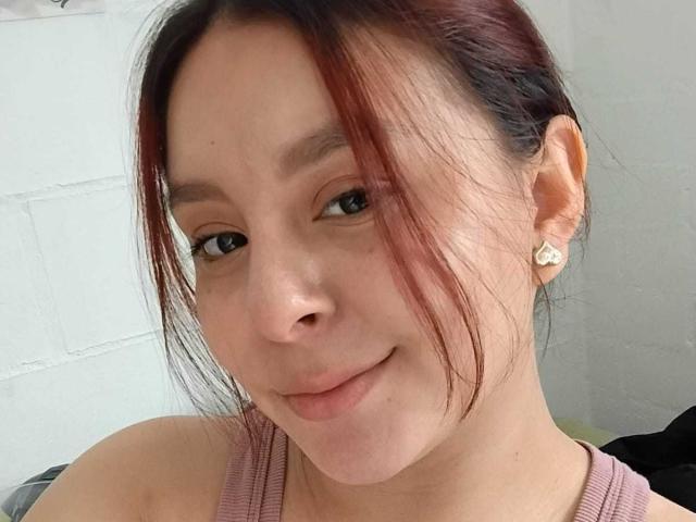 EmaJames with light skin, red-brown hair in a messy bun, small nose, subtle smile, wearing pink tank top, and pearl earrings, looking at camera.