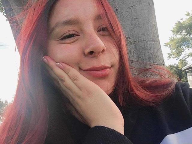 EmaJames with red hair, fair skin, and pink lips, smiling slightly, resting her hand on her cheek. She's wearing a black shirt, standing next to a tree.