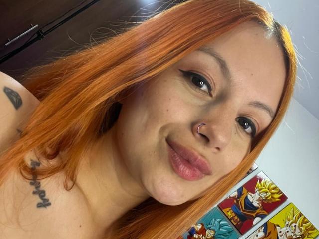 MiiaJonnes with orange hair, black eyeliner, nose ring, and pink lipstick, showing a tattoo on her shoulder, smiling at the camera. Her tits are partially visible.
