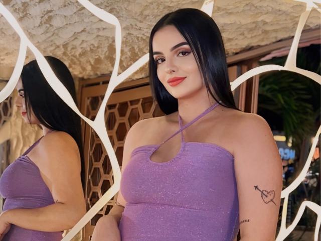 NovaDixon mit langen, schwarzen Haaren, einem violetten, glitzernden Halstopp, rotem Lippenstift und einem Tattoo am rechten Arm, steht vor einem Spiegel, wobei die Spiegelung eine Seitenansicht ihrer Brüste zeigt..