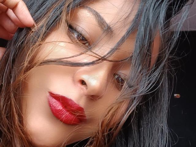 BellaChaudAmour's face, with messy black-gray hair, brown eyes, and shiny red lips, slightly tilted. Her hand touches her head, exposing her nose. She's looking at the camera.