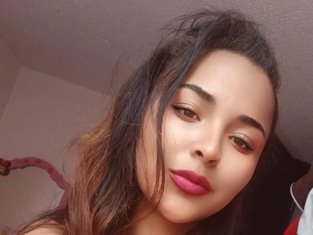 BellaChaudAmour has medium brown skin, dark wavy hair, and pink lipstick. Her eyebrows are thick and she's looking slightly to the side with a relaxed expression.