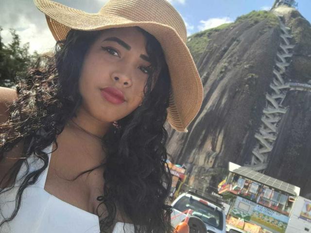 LixAmapola with curly black hair, tan skin, wearing a white tank top and straw hat, smiling, showing her boobs, in front of a tall rock mountain and a car.