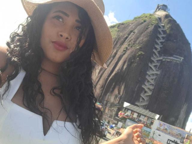 LixAmapola with curly black hair, wearing a white tank top and straw hat, has full tits and a cute face, standing near a tall, rock-climbing structure on a sunny day.