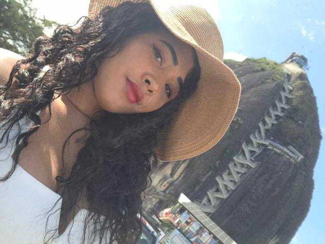 LixAmapola with wavy black hair, tan skin, red lipstick, wearing a white top and tan hat, standing in front of a large rock formation with a bridge.