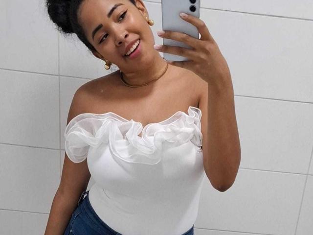 LixAmapola with dark curly hair, smiling, wearing a white off-shoulder top with ruffle trim, blue jeans, gold earrings, and a gold necklace, taking a selfie with a white phone.