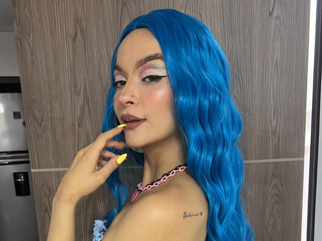 MaryMilan with bright blue wavy hair, yellow nails, pink chain necklace, and bold eyeliner, smirking, touching her lip, tattoo on right shoulder.