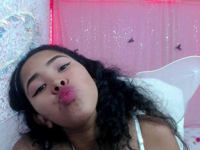 LixAmapola with curly black hair, pouty pink lips, and thick eyebrows, kissing the air, wearing a white top, in a pink, sparkly room.