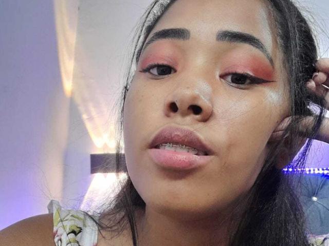 LixAmapola's face, with pink eyeshadow, thick eyebrows, and slightly parted pink lips, holding her hair. She has a light brown skin tone and a subtle sheen on her face.