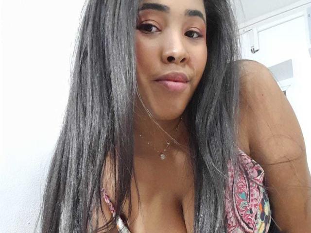 LixAmapola has long, straight black hair, brown eyes, pink lipstick, and a small necklace. She's wearing a colorful, low-cut top showing her large, rounded tits.