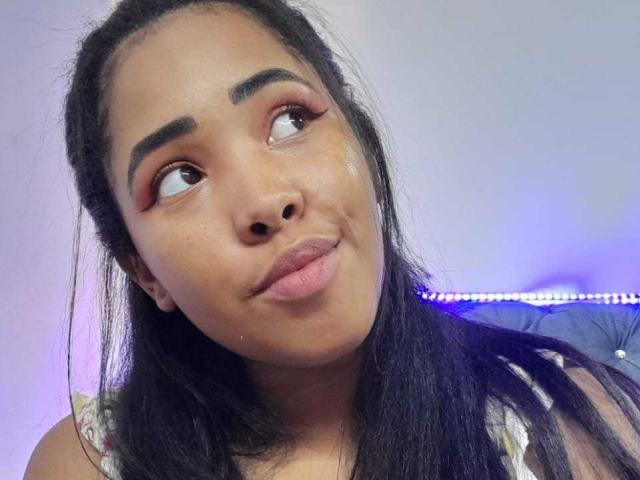 LixAmapola with dark brown eyes, thick eyebrows, and pink lipstick, looking to the side with a slight pout, black hair, and a shiny face, wearing a white top.