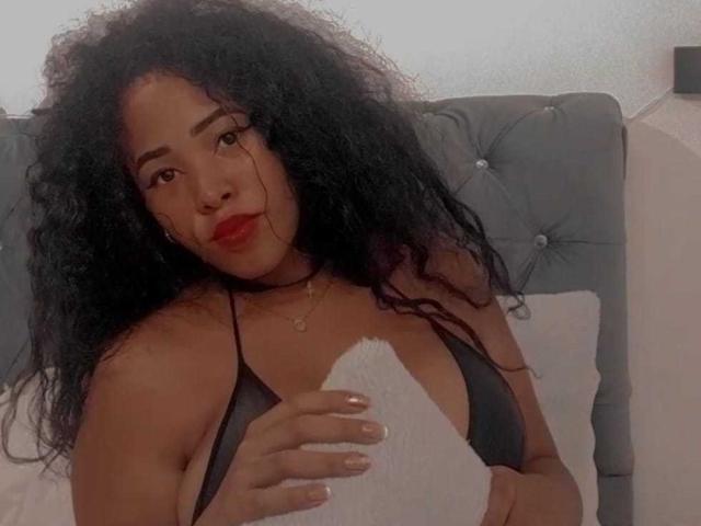 LixAmapola with curly black hair, red lipstick, black bikini top, holding white pillow, grey tufted headboard, looking at camera, soft lighting, slight smile. Boobs visible, brown skin.