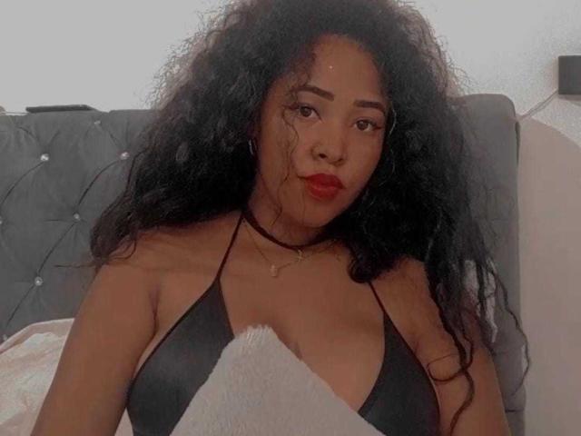 LixAmapola with curly black hair, wearing a black bikini top, showing her tits, red lipstick, black choker, and a fuzzy white blanket in a soft, dimly lit setting.