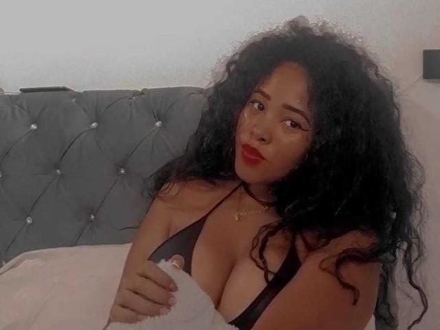 LixAmapola with curly black hair, red lipstick, and black bikini top, showing large tits, holding a white blanket in a tufted grey headboard bedroom.