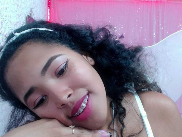 LixAmapola with curly black hair, white headband, pink eyeliner, and pink lipstick, smiling with teeth showing, resting her chin on her hand, wearing a white strap top.