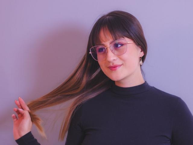NinaBryons with long brown hair, holding a lock, wearing glasses and a black turtleneck, smiling with pink lipstick, fair skin, small earrings, in a purple-lit photo.