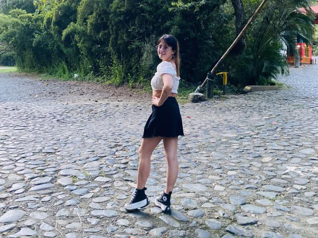 NinaBryons stands on a cobblestone path, wearing a white cropped top, black skirt, and black sneakers with white soles, looking back with a slight smile.