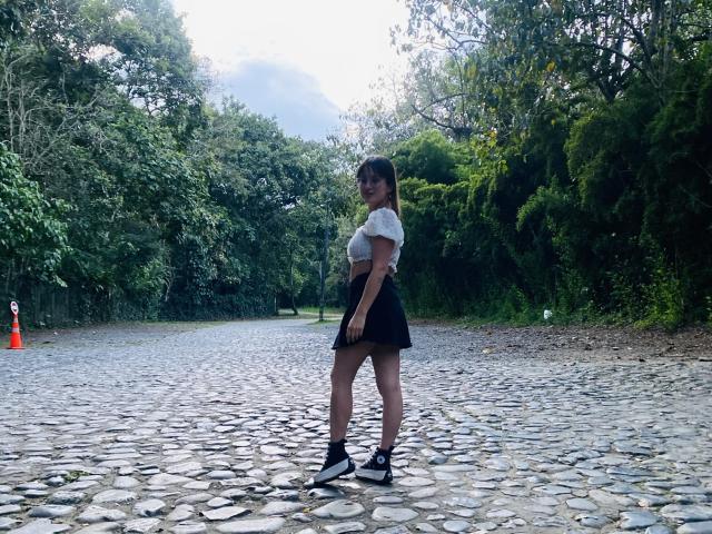 NinaBryons standing on a cobblestone path, wearing a white crop top, black skirt, black socks, and black sneakers, with her back to the camera, looking over her shoulder.