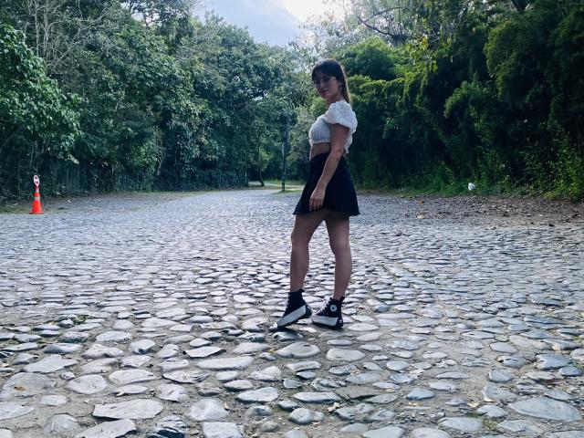 NinaBryons with a white t-shirt, black skirt, and black sneakers, standing on a cobblestone path, showing her ass and looking back with a smile.