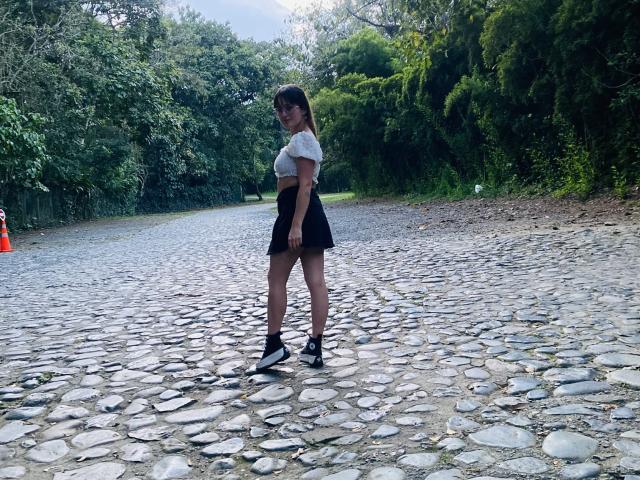 NinaBryons stands on a cobblestone path, wearing a white shirt, black skirt, and black sneakers, with her ass facing the camera, looking back over her shoulder.