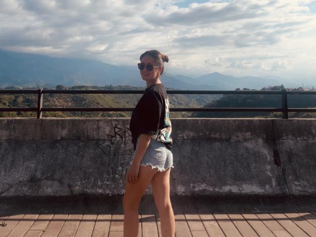 NinaBryons stands on a brick patio, facing sideways with a smile. She's wearing sunglasses, a black shirt, and frayed denim shorts, showing her ass. Mountain view in background.
