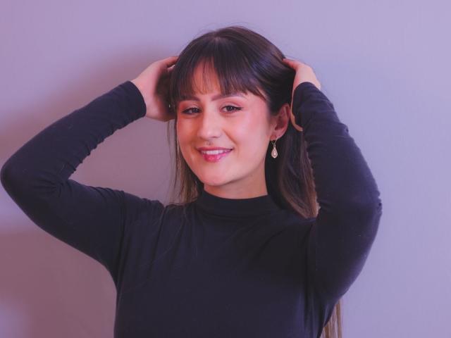 NinaBryons with hands in dark brown hair, smiling, wearing a black long-sleeve top, small hoop earrings, pink lipstick, and a nose ring, against a plain background.
