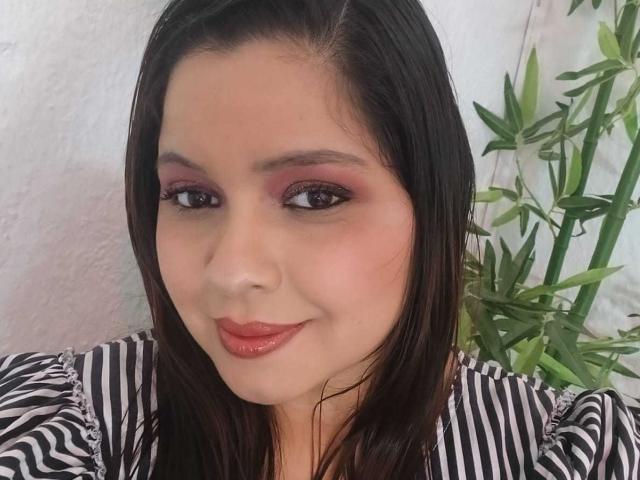 Idylla Jhones, con capelli castano scuro, ombretto rosa, rossetto rosso e una camicia a righe, sorride con le labbra lucide, guardando la fotocamera, con una pianta di bambù a destra..
