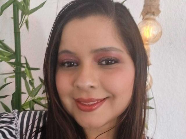 IdyllaJhones has brown hair, red lipstick, and pink eyeshadow, smiling with a cute face, medium tits, and a warm light bulb in the background.