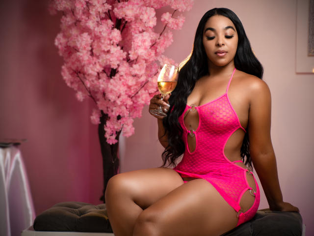 EileenBenzon in pink, cut-out lingerie, holding a champagne glass, with long black hair, sitting on a bench, showing her curvy ass and small, perky tits.