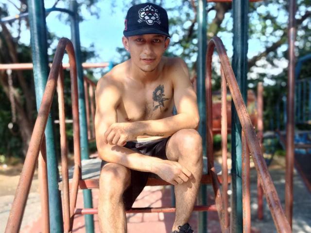 Shirtless JacobHuges, with a tattoo on his chest, wearing a black cap and black shorts, sits on a rusty metal playground structure, arms crossed, legs crossed, looking down.