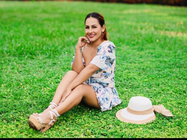 BombSophie with red hair, smiling, sitting on grass, wearing floral dress, white sandals, hand on chin, white hat and beige ribbon on ground, legs crossed, red lipstick.