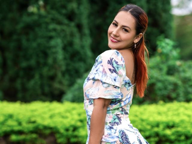 BombSophie with brown hair, smiling, wearing a floral dress, showing her ass, green shrubbery in background, earrings, looking over shoulder, red lipstick, white dress with blue pattern.