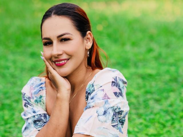 BombSophie with brown hair, smiling, hands near face, wearing floral shirt, red lipstick, gold earrings, green grass background, looking cute and sexy, soft light, casual pose.