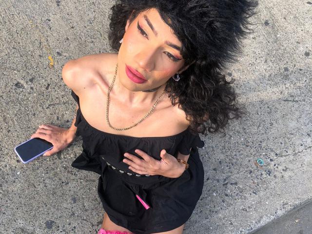 SweetSamyPrincipal with curly black hair, wearing an off-shoulder black dress, holding a phone, gold chain, pink lipstick, looking up, hands on chest, standing on a rough concrete surface.