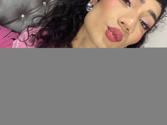SweetSamyPrincipal with curly black hair, pink lipstick, and green earrings, pouting her plump, glossy lips. Wearing a pink shirt, she's got a cute, sexy look.