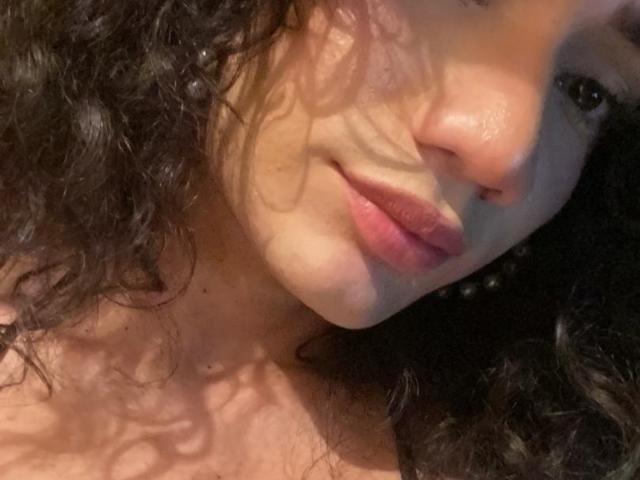 SweetSamyPrincipal's face, showing curly black hair, pinkish lips, and a slight smile, with soft shadows on her cheek and neck, in a slightly angled, close-up selfie.